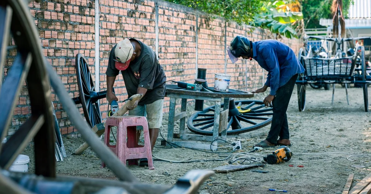 Deux artisans travaillent à la restauration de vieilles roues de calèche en plein air, à l'aide d'outils, entourés de verdure.
